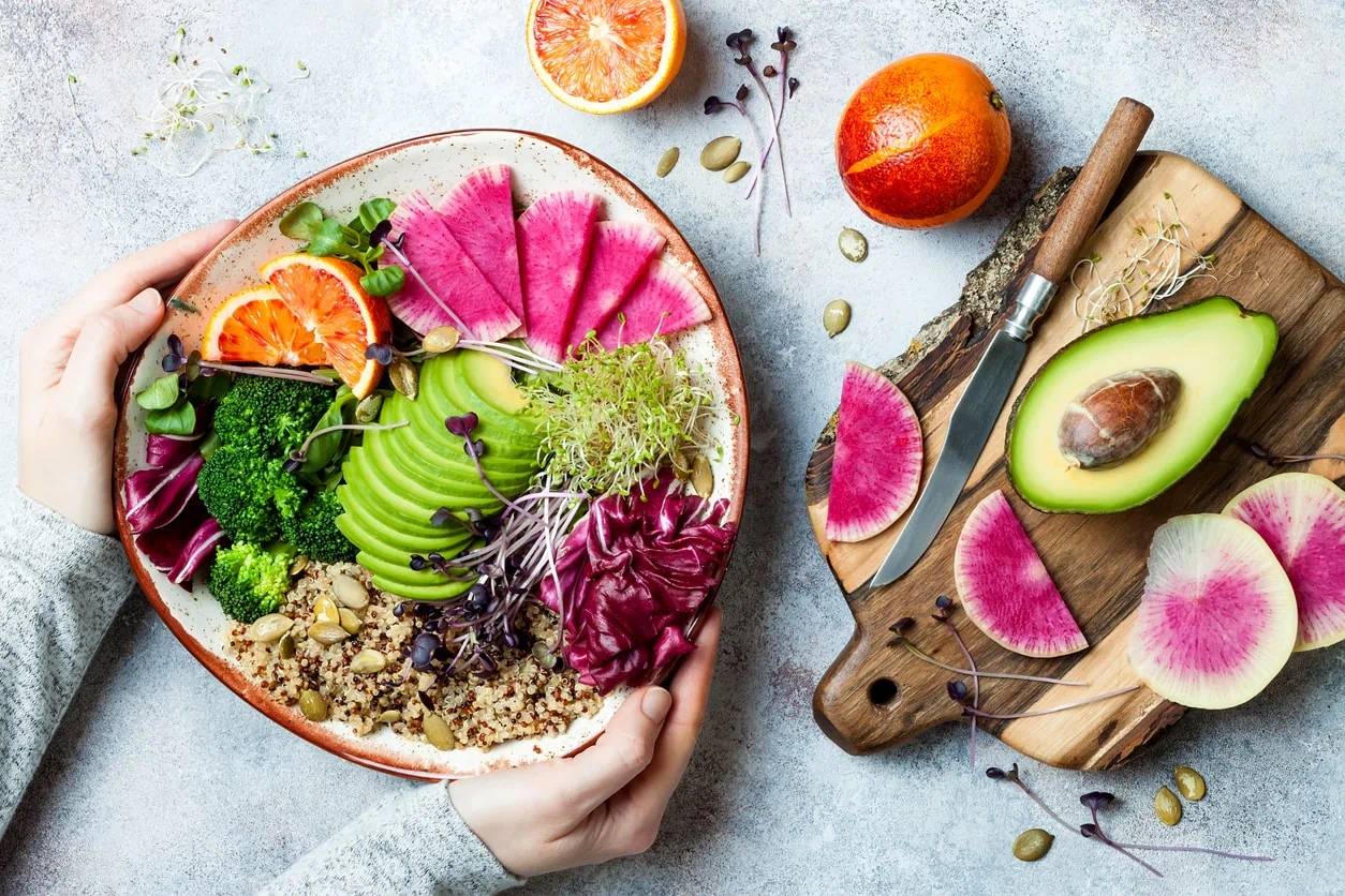Hands holding vegan, detox Buddha bowl with quinoa, greens, avocado, oranges, broccoli, watermelon radish, alfalfa seed sprouts.