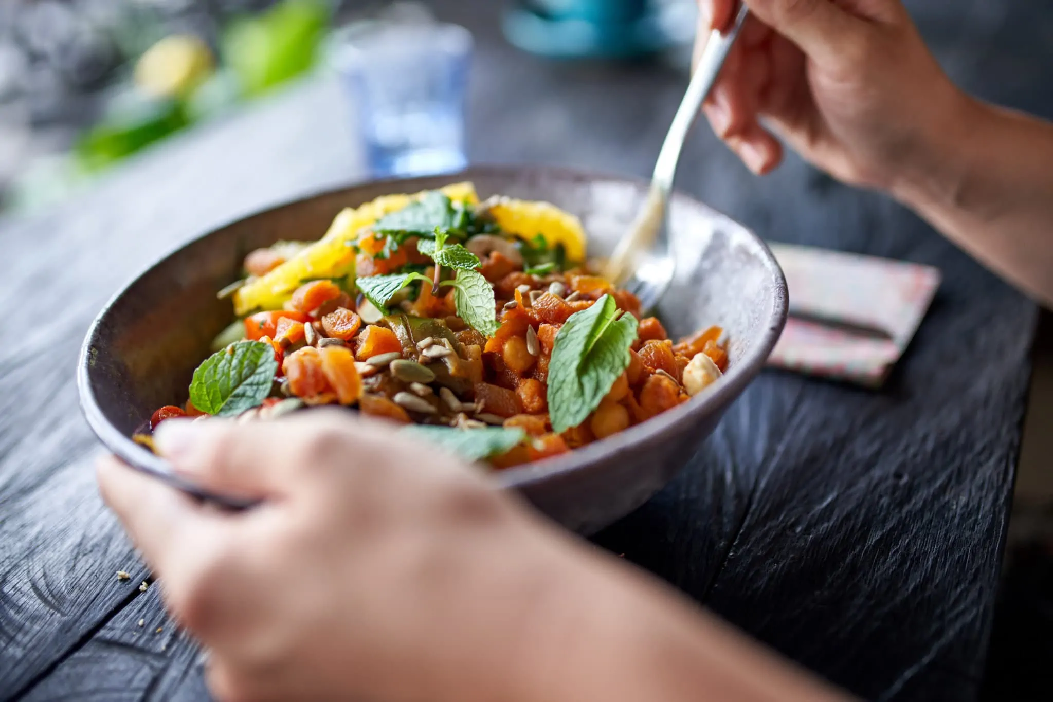 Closeup shot of hands holding a bowl with chickpeas, fresh herbs and fruit at wooden table