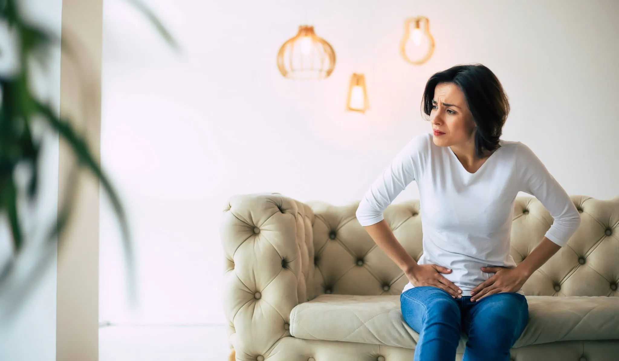 Young woman is sitting on a sofa and touching her lower stomach in discomfort.